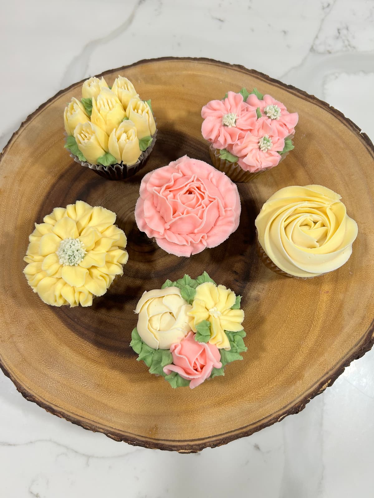 Five floral buttercream cupcakes arranged on a wood slice โ the Sip and Pipe workshop